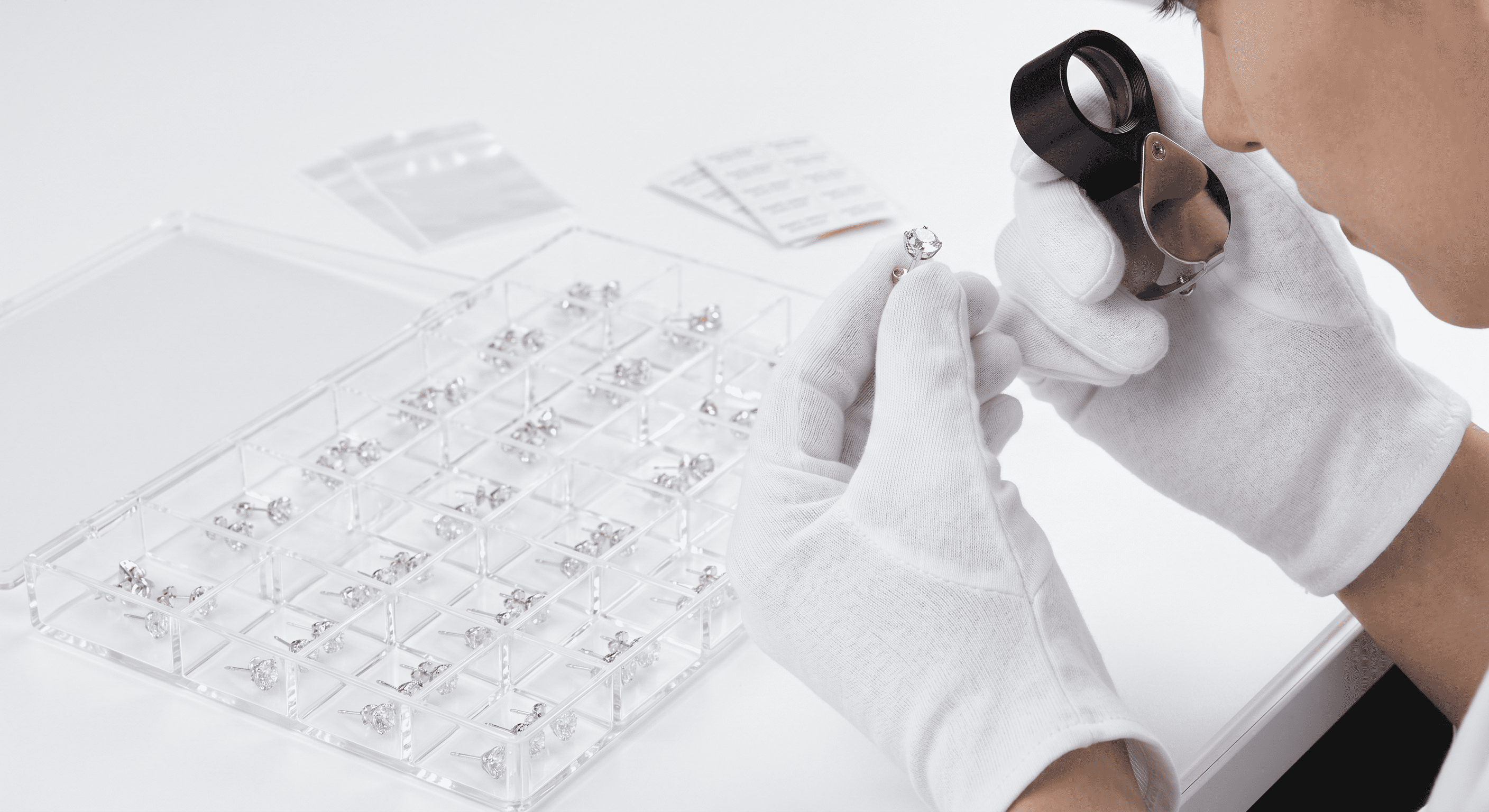 A quality control technician wearing immaculate white cotton gloves holds a silver stud earring up to a professional optical jeweler's loupe (1) A quality control technician wearing immaculate white cotton gloves holds a silver stud earring up to a professional optical jeweler's loupe (1)
